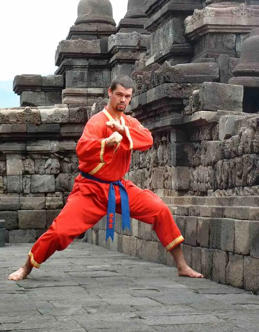 Een Tapak Suci-beoefenaar in rood pak toont een lage gevechtshouding bij een stenen tempel.
