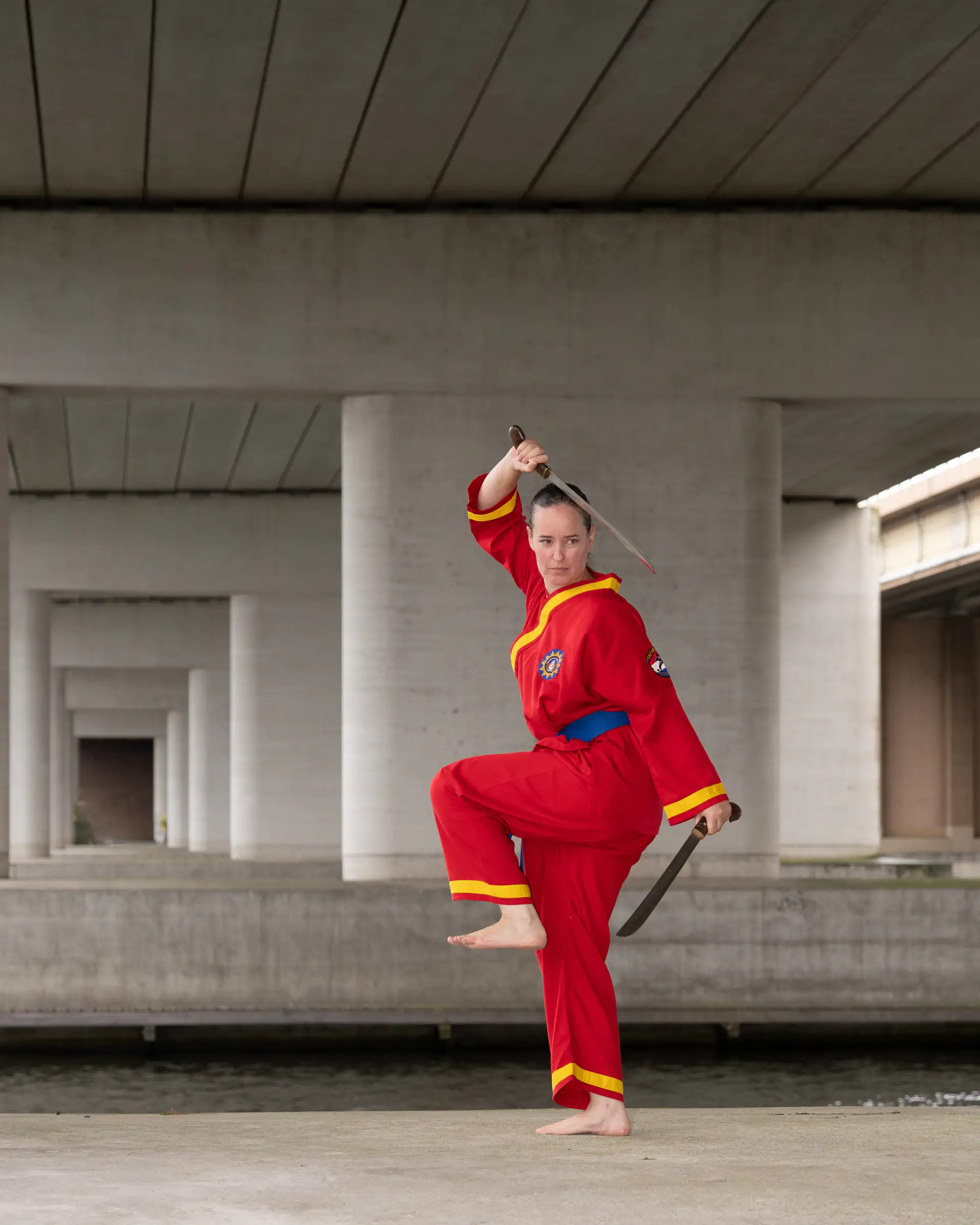 Een Tapak Suci-beoefenaar in een rood pak balanceert op één been met twee trainingswapens onder een brug.